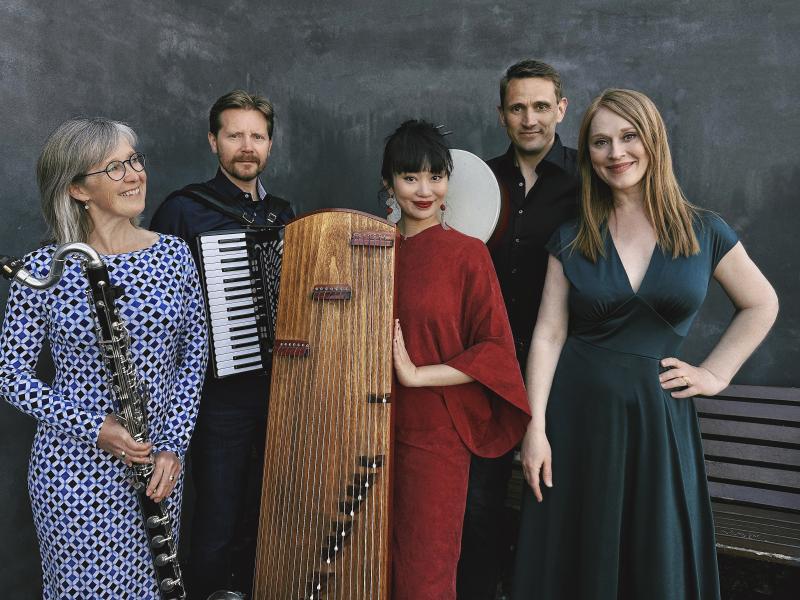 Fem velklædte musikere ved siden af hinanden, i midten står en kvinde med et kinesisk musikinstrument, en guzheng. 